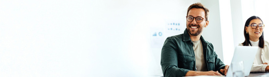 Selbstständiger Mann sitzt an seinem Laptop, im Hintergrund eine Frau.