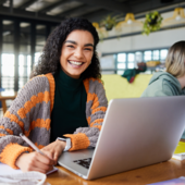 Studentin sitzt vorm Laptop und lacht in die Kamera.